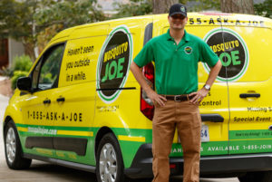 Mosquito Joe technician in green short sleeved shirt and khaki pants posing for photo behind black and yellow service van.
